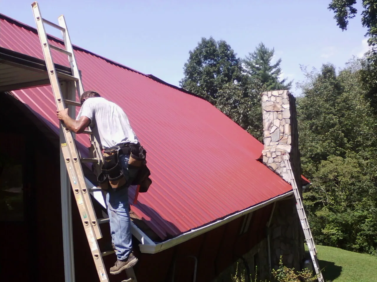 Roofer on a ladder beside a red metal roof and stone chimney — Soffit & Fascia Repair by Roofing Brothers in Manito
